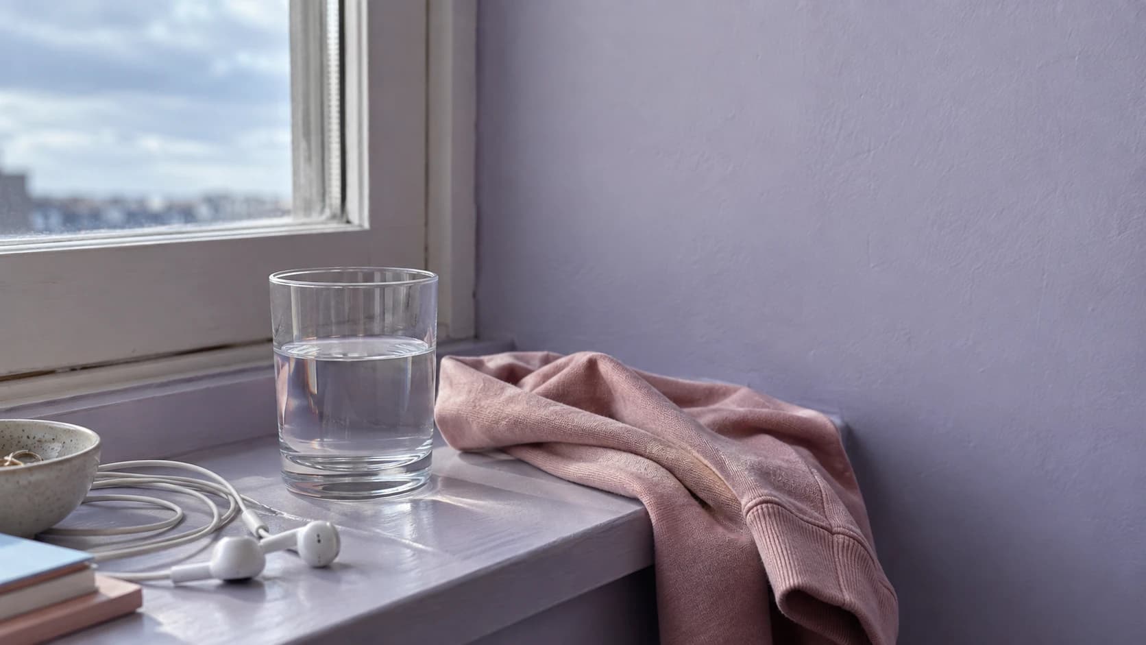 A glass of water and earbuds on a windowsill beside a soft rose sweater.