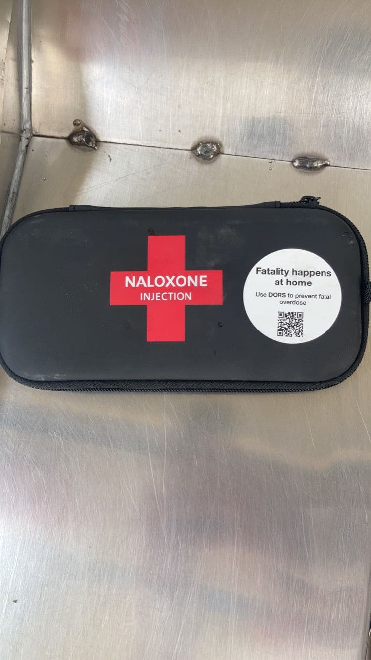 A black zip-up naloxone case with a red cross and the word NALOXONE on the front, sitting on a metal surface